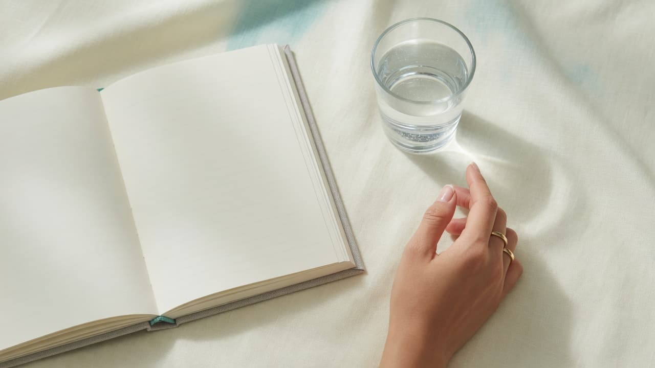 Calm hand with natural nails beside an open journal and glass of water on pale linen