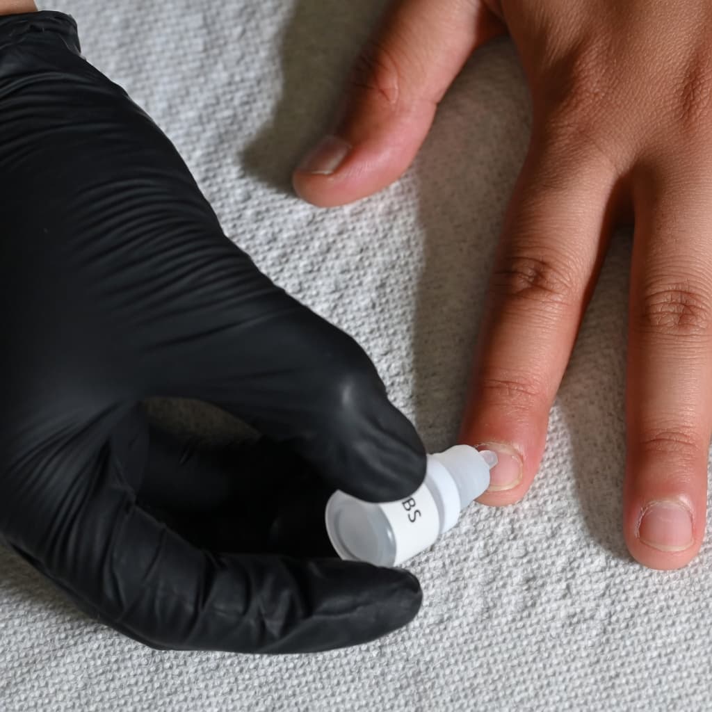 Gloved hand applying a small amount of nail glue onto a natural fingernail